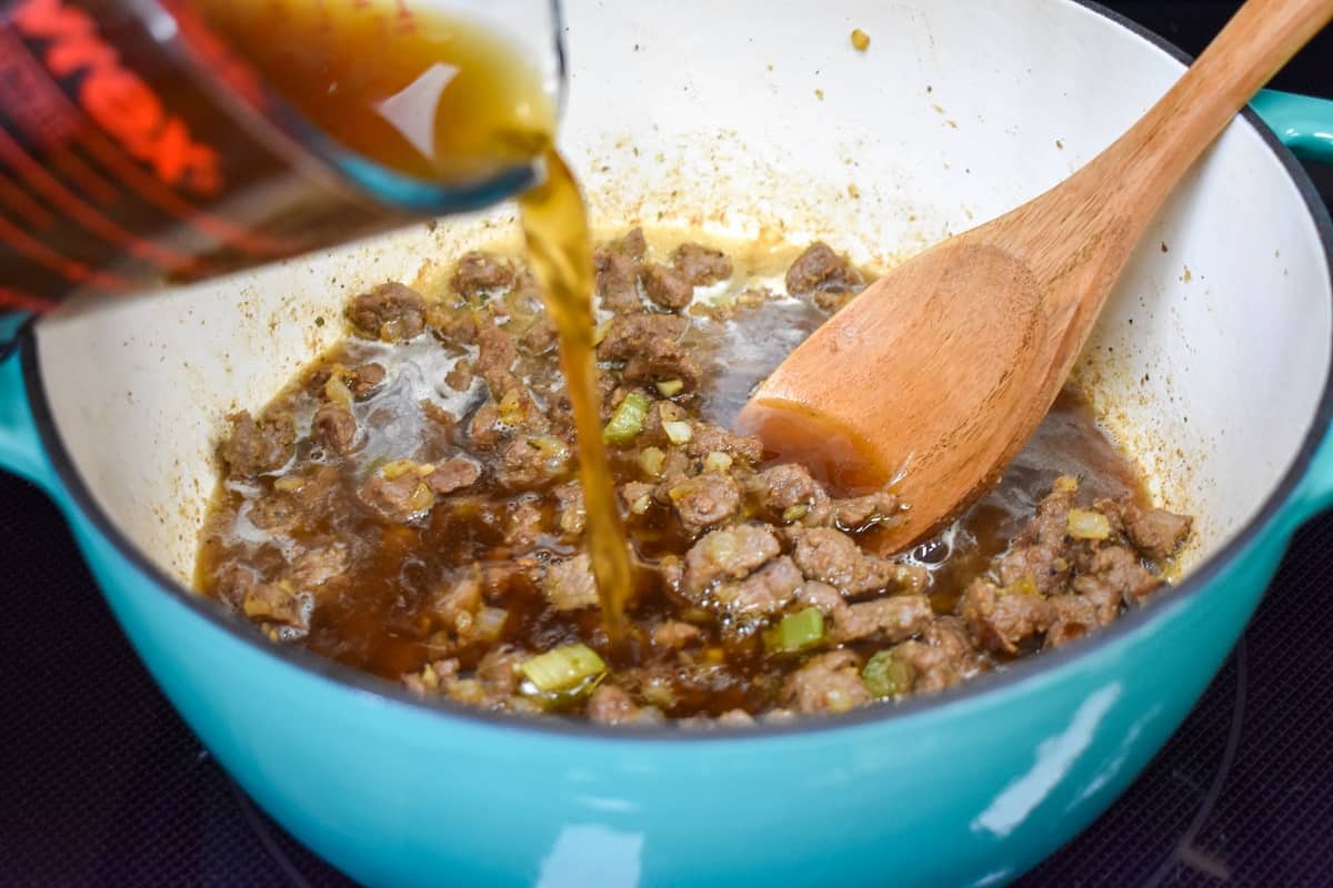 Beef broth being added with a measuring cup to the beef and vegetable mixture in the pot.