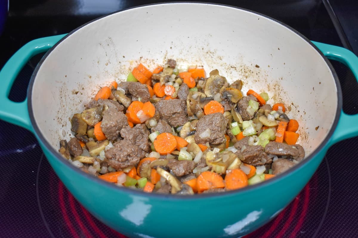 Browned beef pieces with mushrooms, carrots, onions, and celery cooking in a large pot.