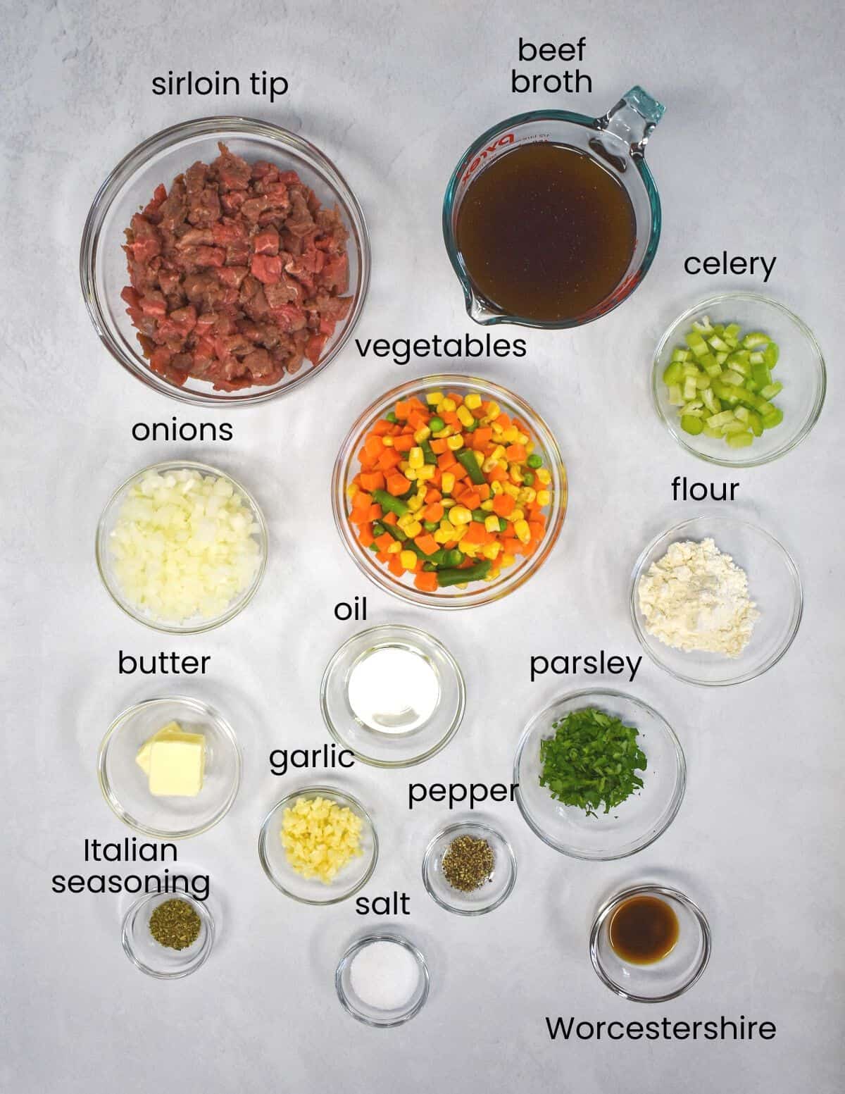 The ingredients for the pot pies arranged in glass bowls on a white table, with each ingredient labeled with black letters.