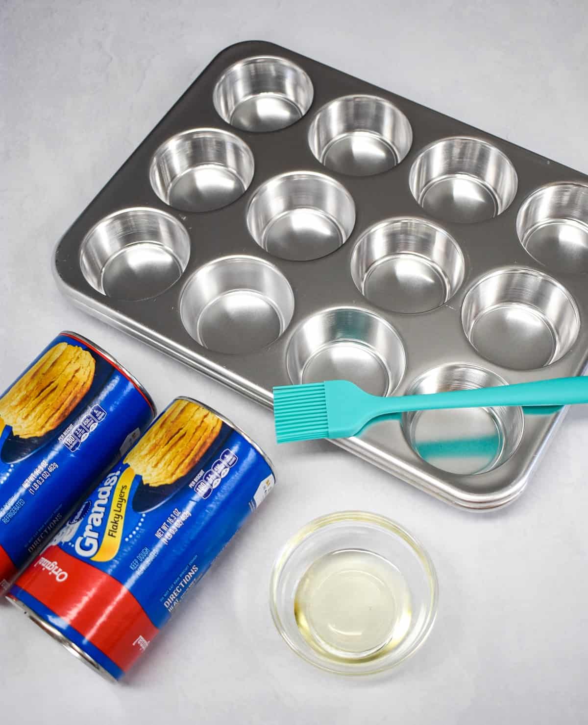 Two packages of refrigerated biscuits, muffin pan, oil in a small glass bowl, and a pastry brush.