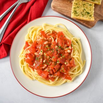 The fresh tomato pasta served on a white plate with a red rim, with a red linen and silver to the left and garlic bread to the top right side.