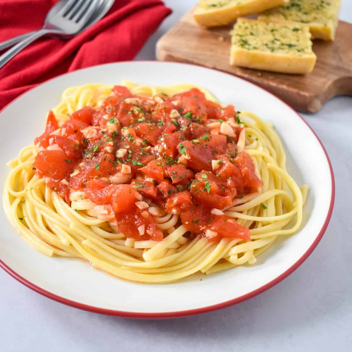 The finished pasta with tomatoes served on a white plate with a red rim, a red linen and silverware to the left side and garlic bread to the top right.