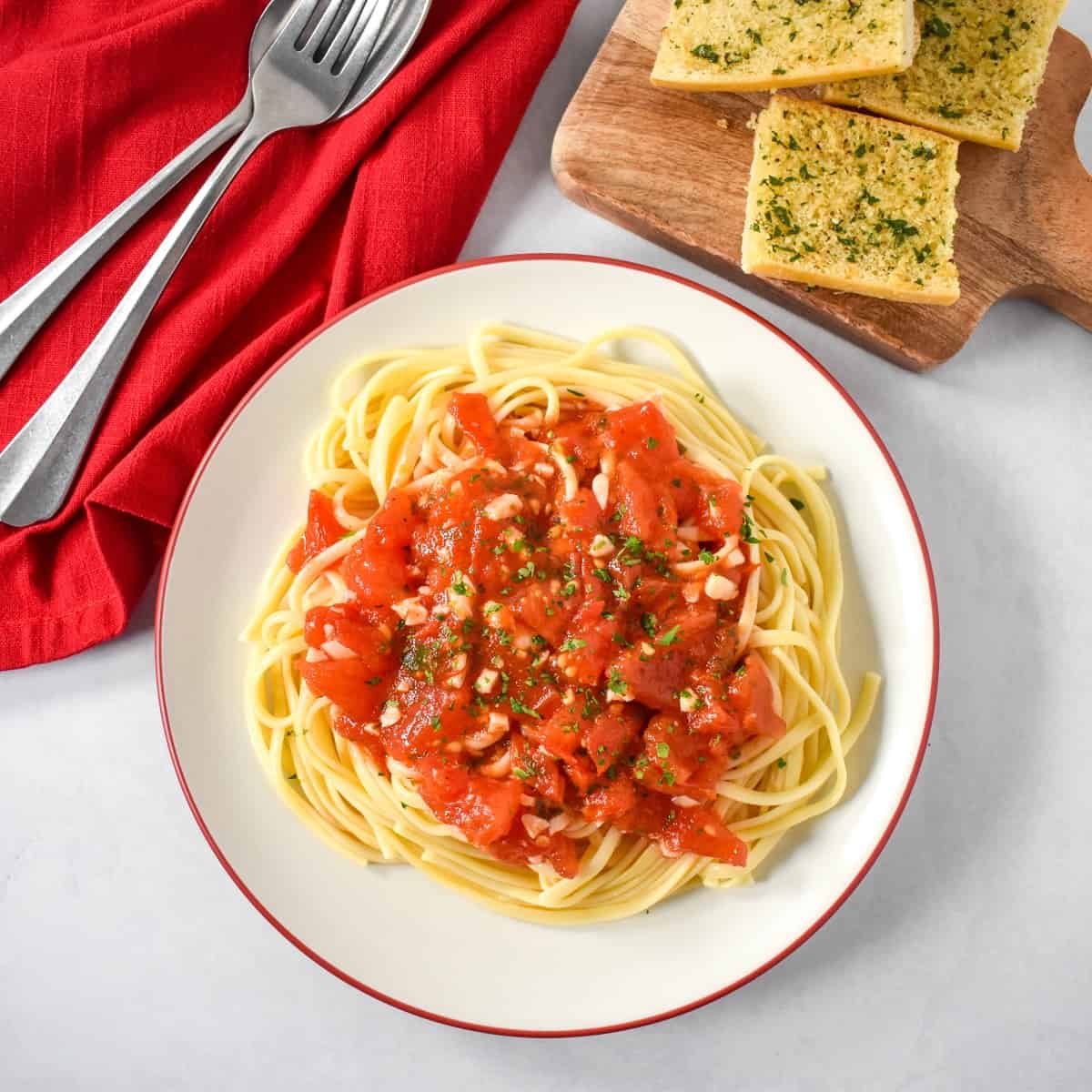 The fresh tomato pasta served on a white plate with a red rim, with a red linen and silver to the left and garlic bread to the top right side.