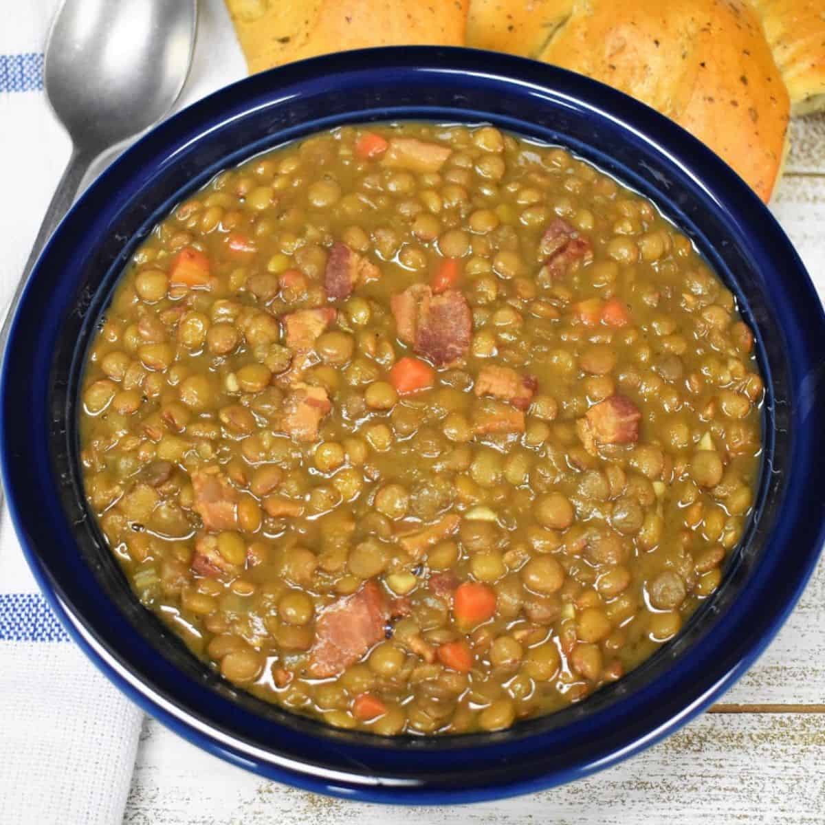 Bacon and lentil soup served in a blue bowl with a piece of bread in the background and a spoon to the left.