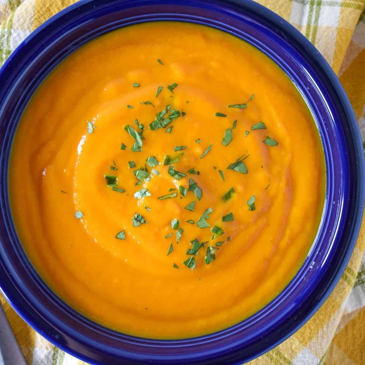 Pureed carrot ginger soup served in a blue bowl on a yellow, green and white checkered linen.