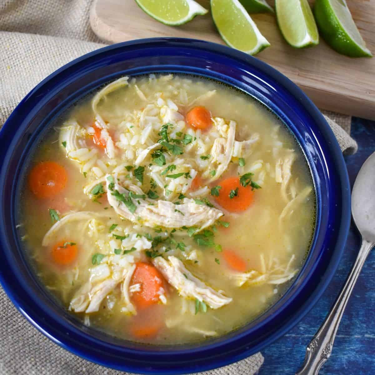 The chicken rice soup served in a blue bowl with lime wedges and hot sauce in the background. All served on a blue table with a beige linen.