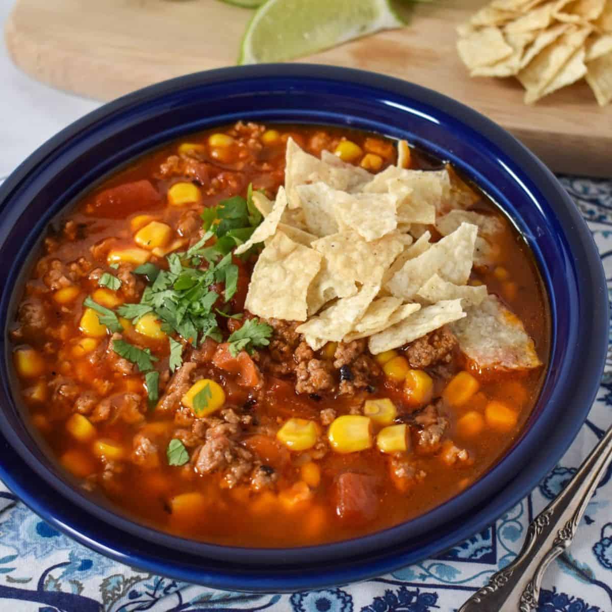 The soup served in a blue plate and displayed on a blue and white linen with lime wedges and extra tortilla chips in the background.