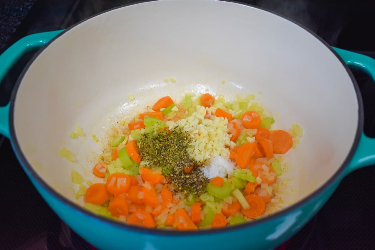 Minced garlic and seasoning added to the carrots, onions, and celery in a large aqua and white pot.