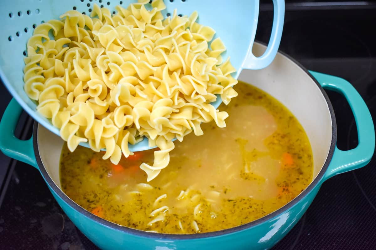 Cooked egg noodles being added to the soup in an aqua and white pot.