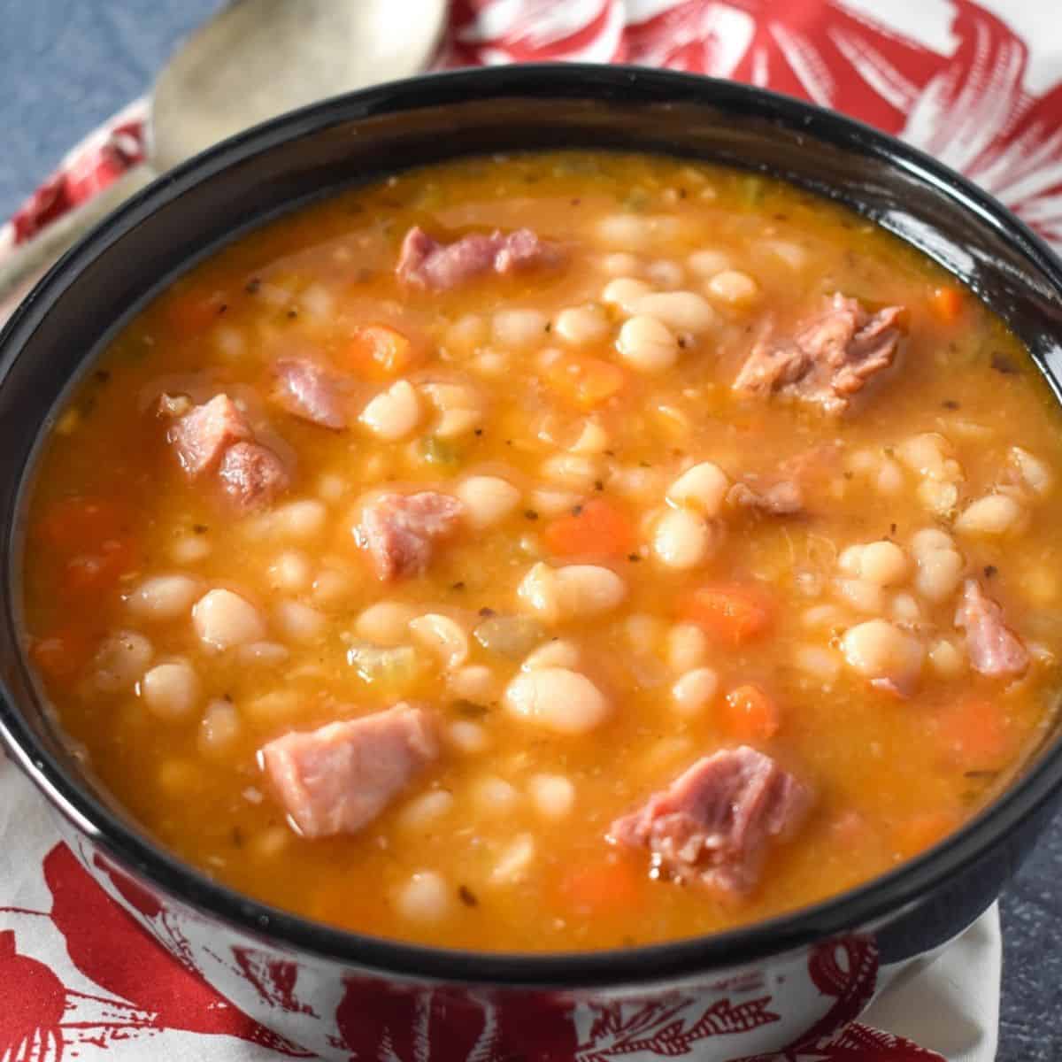 Navy bean soup served in a black bowl, with a red and white cloth napkin and spoon on the side.