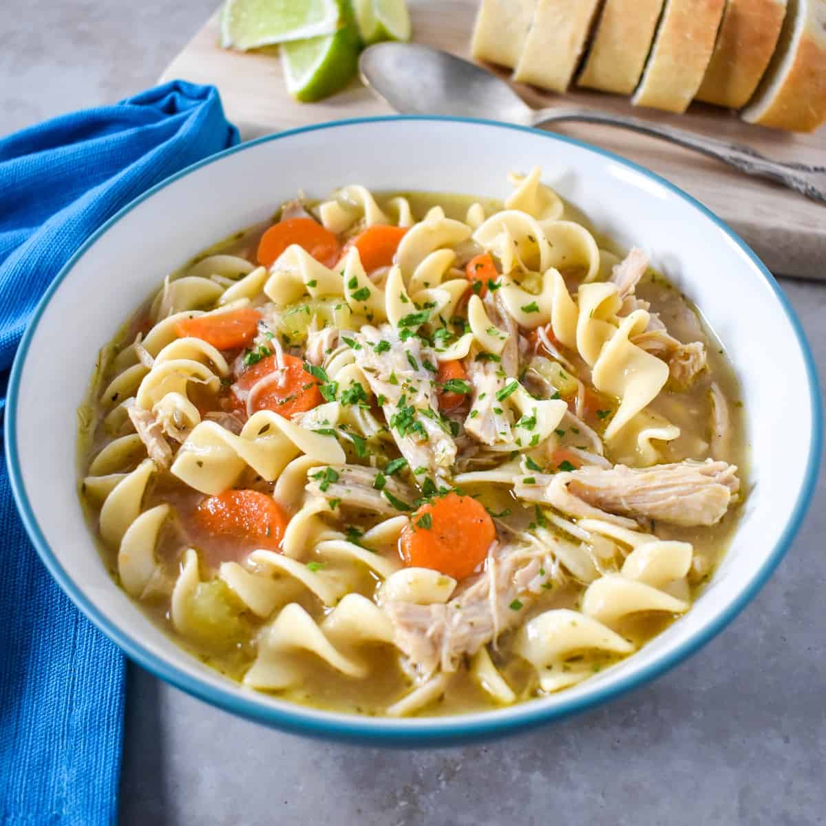 The turkey noodle soup served in a white bowl with bread slices and lime wedges in the background and a blue linen to the left side.