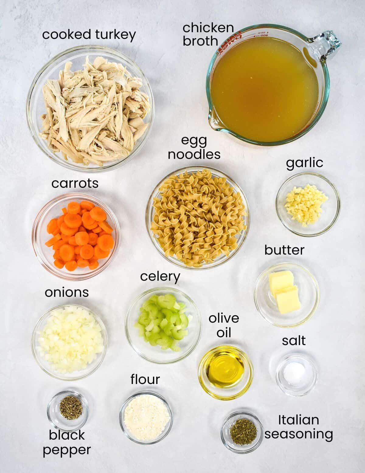 The ingredients for the soup arranged in glass bowls on a white table, each ingredient is labeled in small black letters.