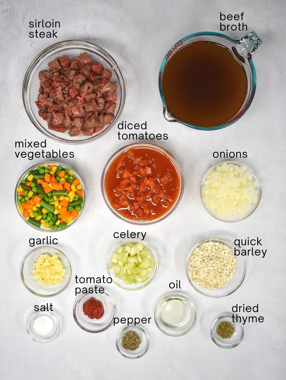 The ingredients for the beef barley soup prepped and arranged in glass bowls on a white table. Each ingredient is labeled with small black letters.