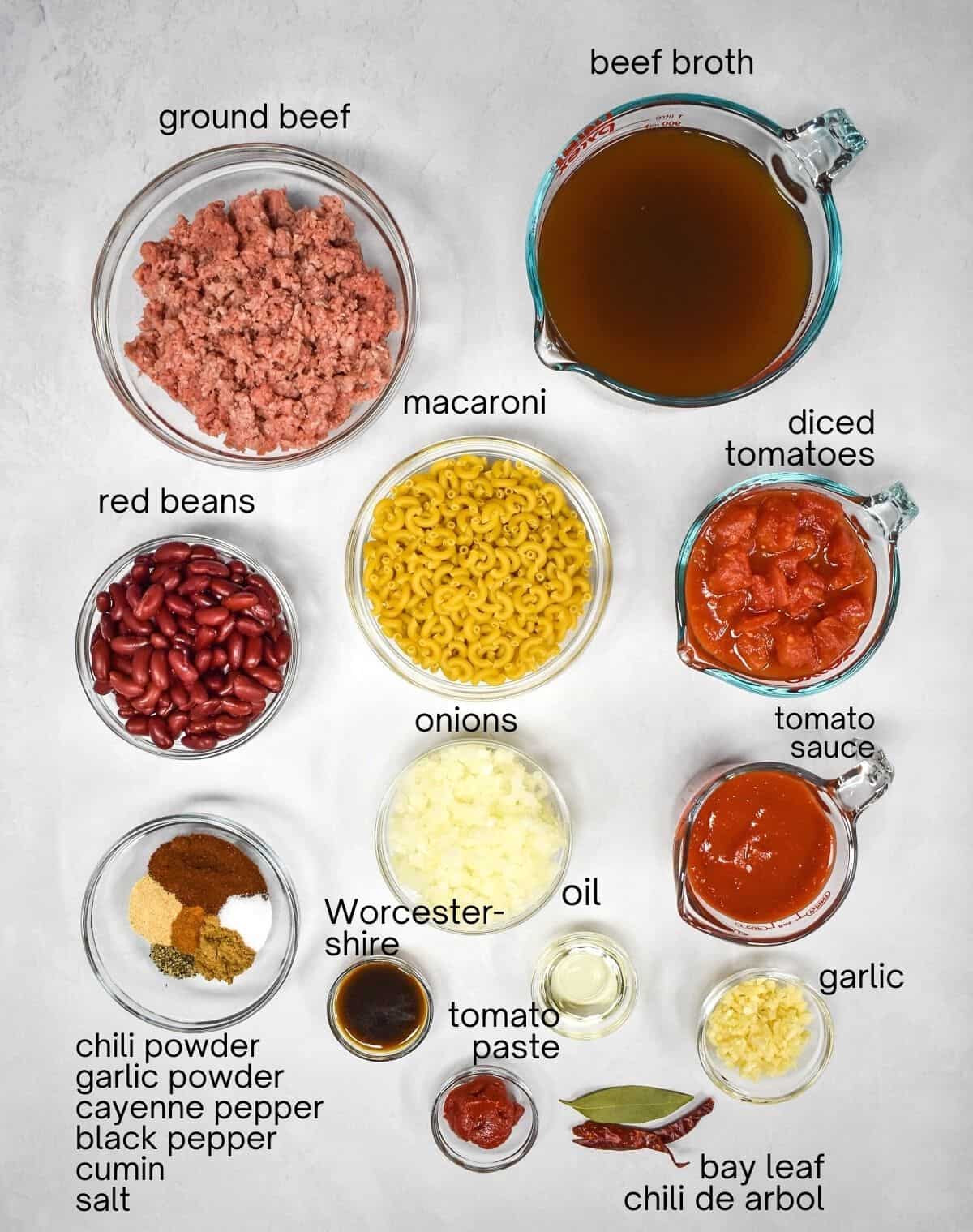 The prepped ingredients for the easy chili mac arrange in glass bowls on a white table. Each ingredient is labeled with small black letters.