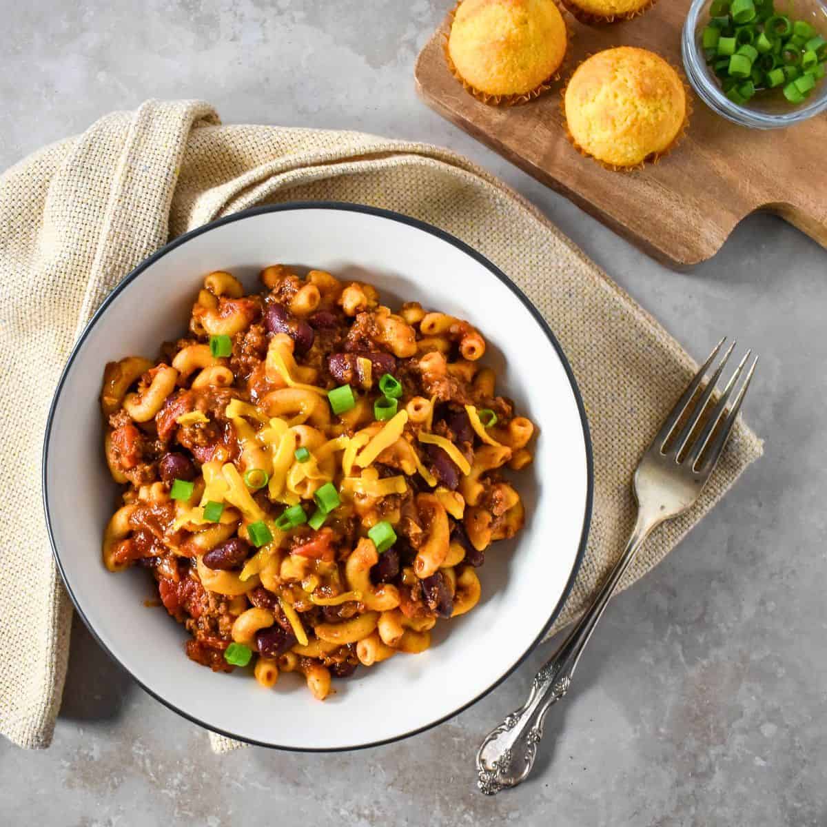 Bowl of easy chili mac with ground beef, kidney beans, and elbow macaroni, topped with shredded cheese and green onions, with cornbread on the side.