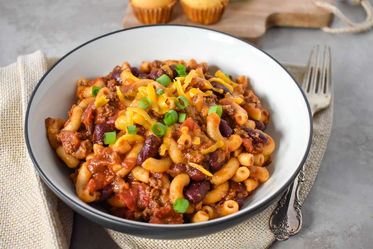 The chili macaroni topped with shredded cheese and sliced green onion served in a white bowl with a black rim.