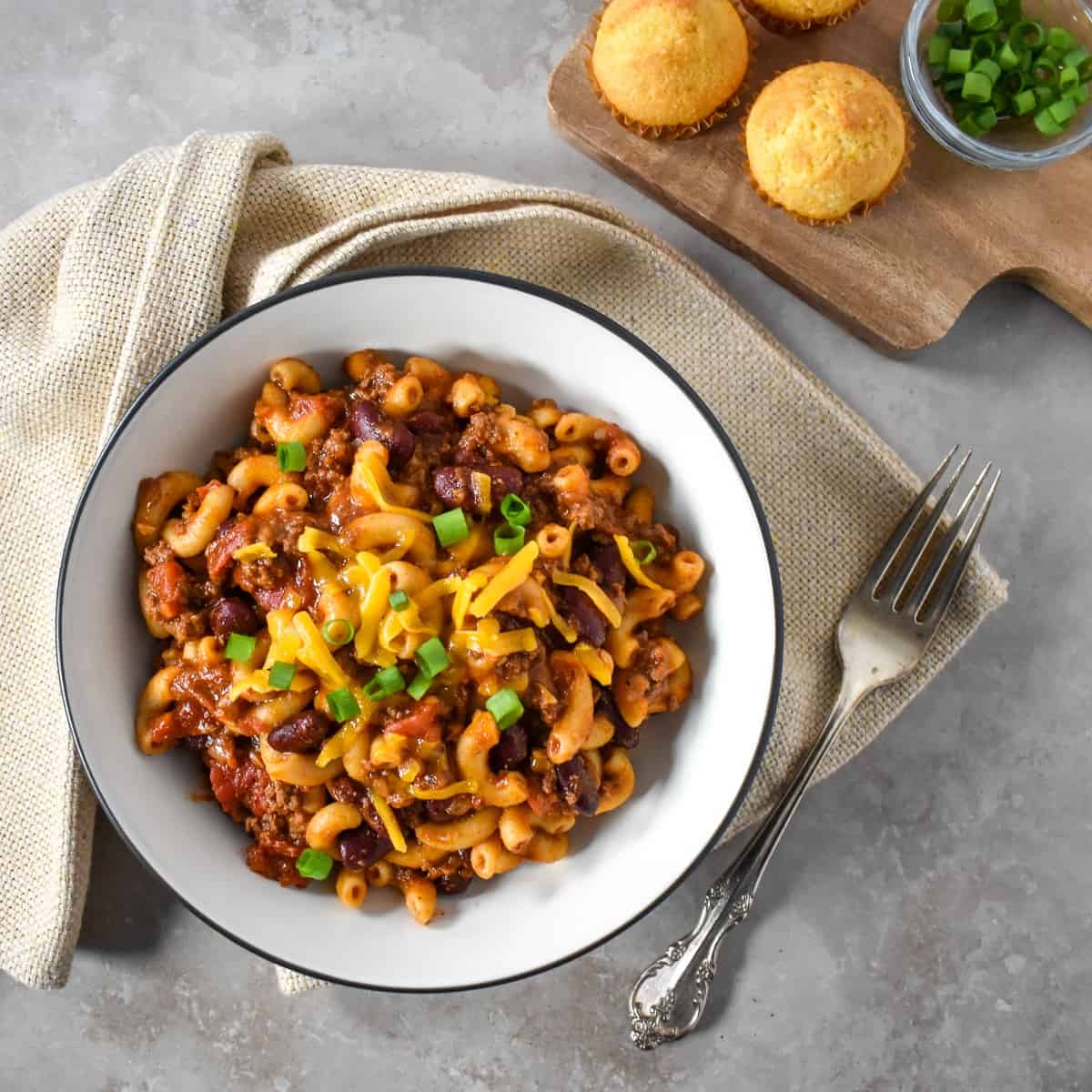 The chili mac served in a white bowl with a black rim, there are mini corn bread muffins and sliced green onions to the top right of the bowl.