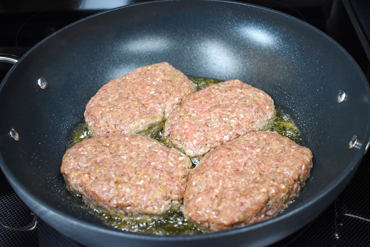 Four raw beef patties frying in a large, non-stick skillet.