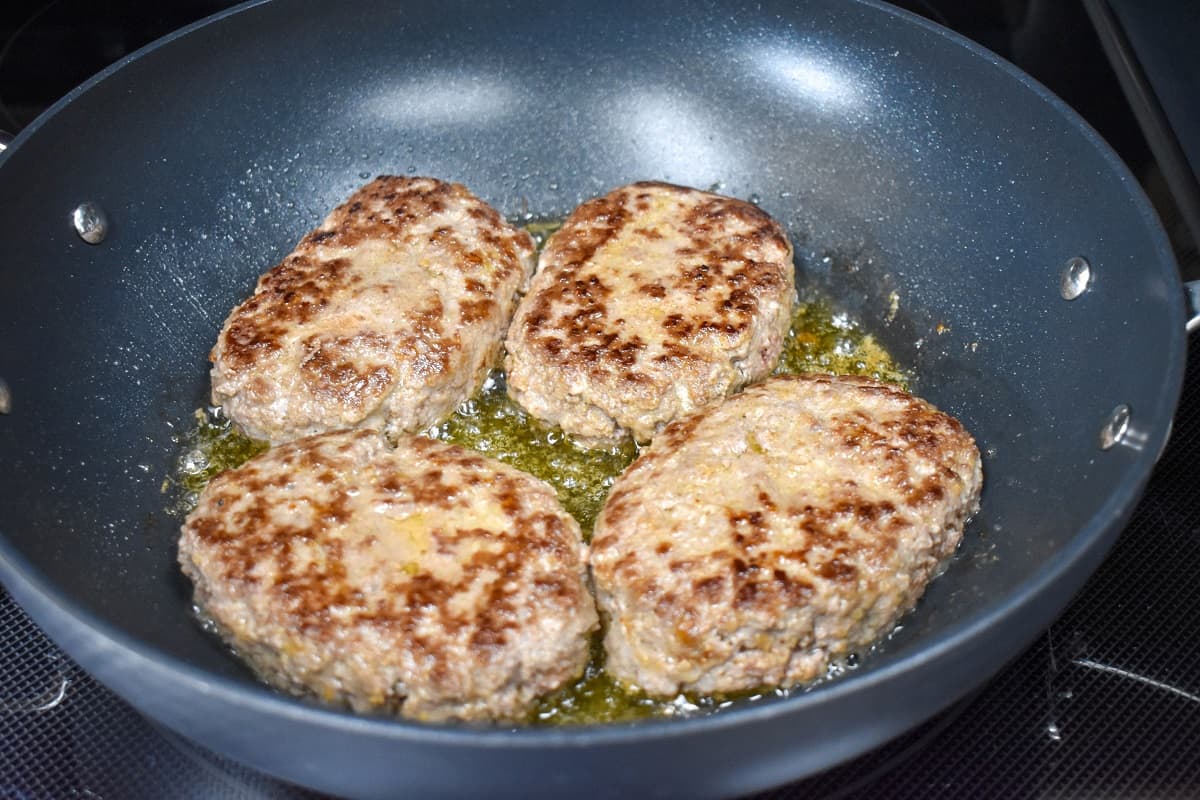 Four browned beef patties frying in a large, non-stick skillet.