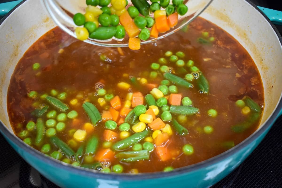 Mixed vegetables being added to the soup in a large pot.