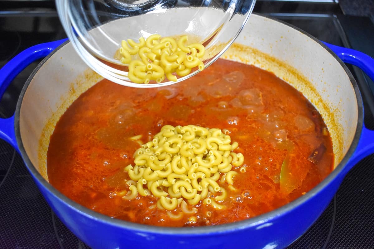 Macaroni being added to the chili in a large white and blue pot.