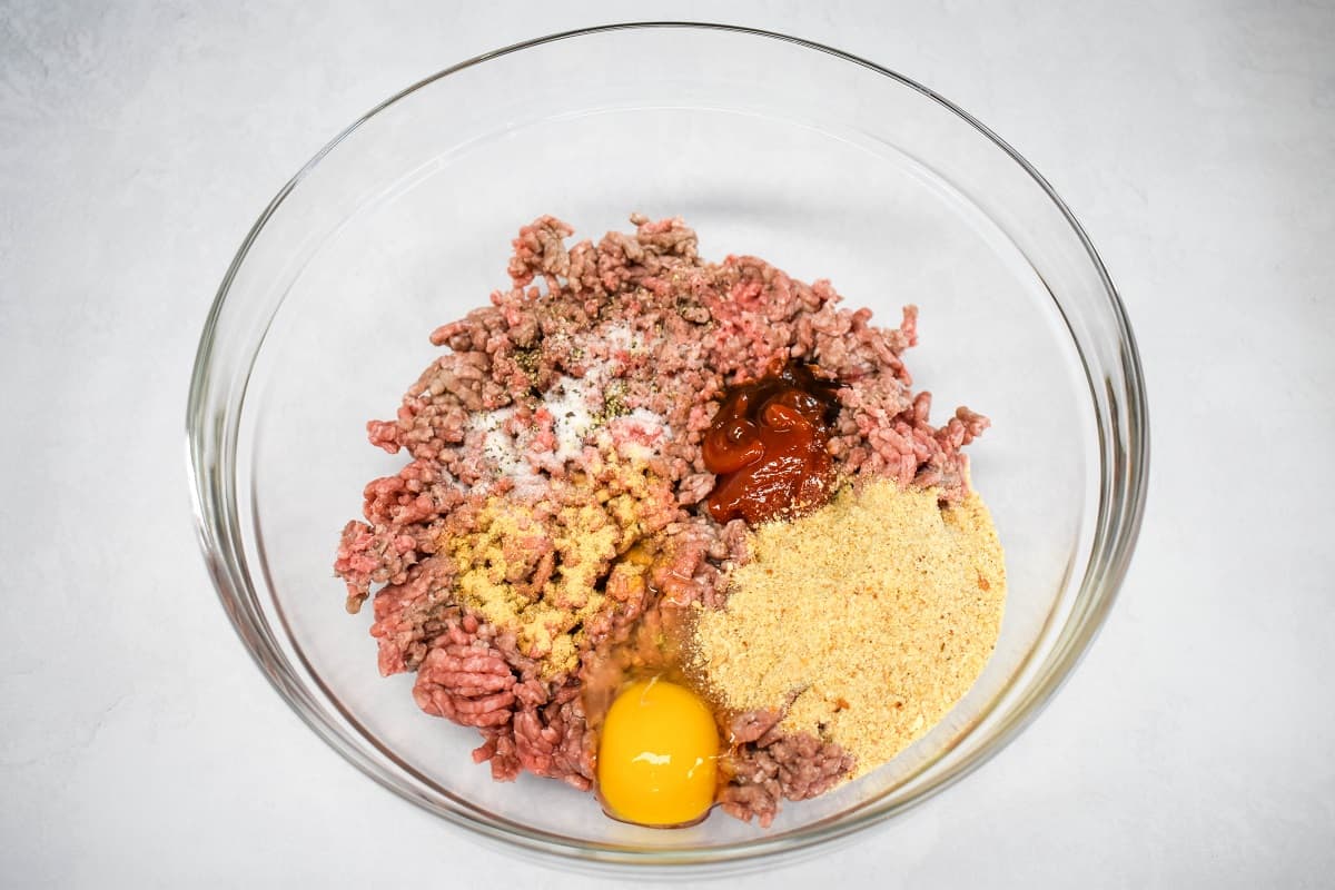 The ground beef, spices, condiments, breadcrumbs, and egg in a large, glass bowl.