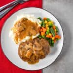 The Salisbury steak served with mashed potatoes and mixed vegetable served on a white plate set on a red linen.