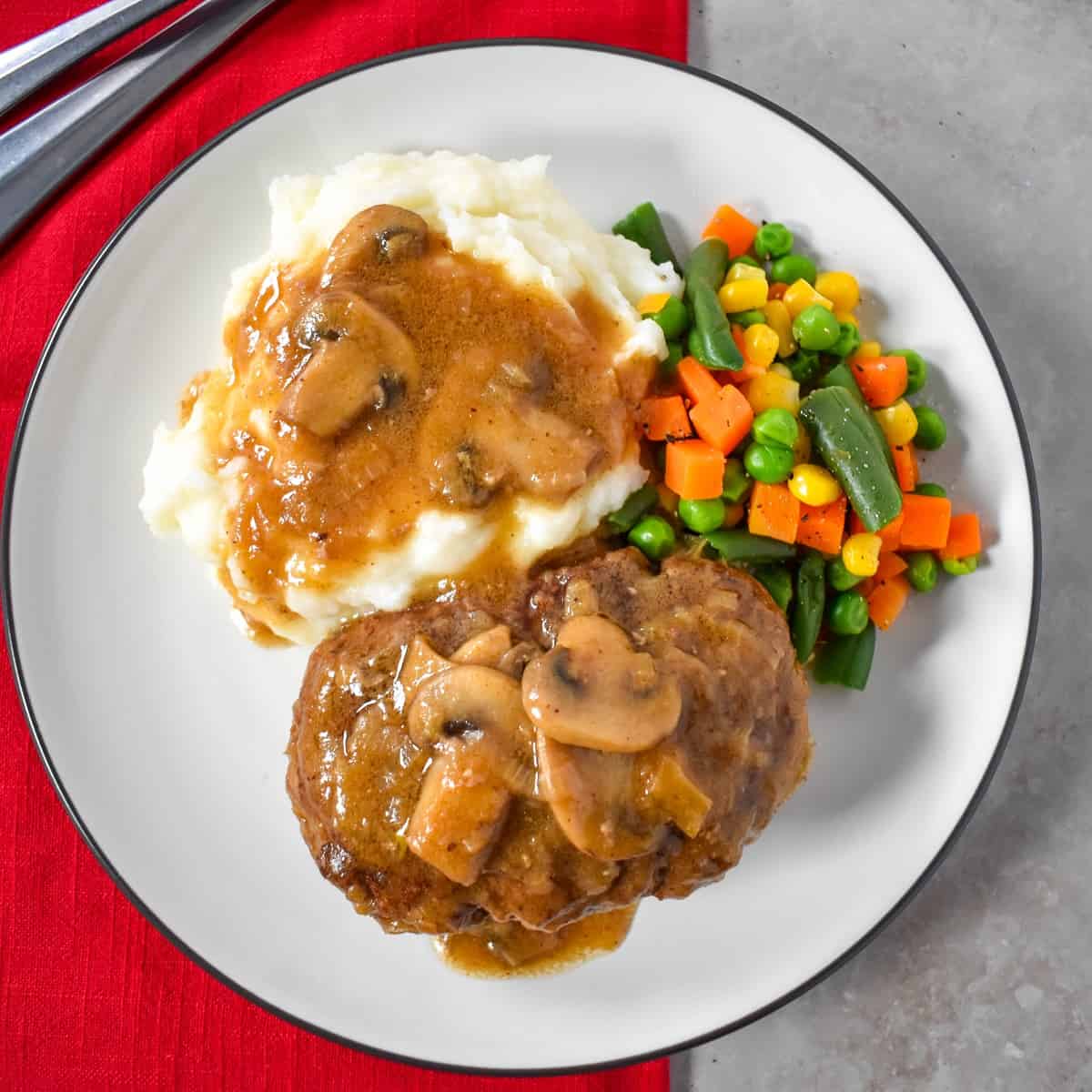 The Salisbury steak served with mashed potatoes and mixed vegetable served on a white plate set on a red linen.