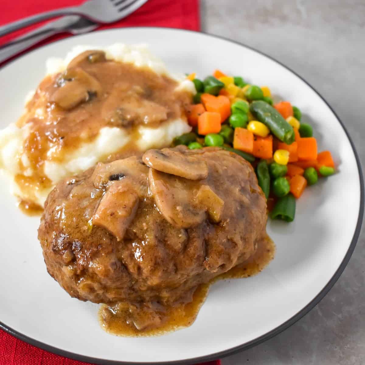 A close up of the Salisbury steak topped with the gravy and served with mashed potatoes and mixed vegetables on a white plate.