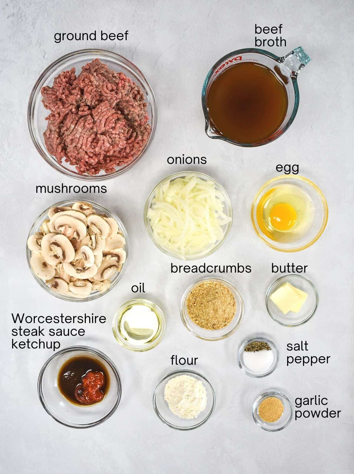 The prepped ingredients for the Salisbury steak arranged in glass bowls on a white table. Each ingredient is labeled with small black letters.