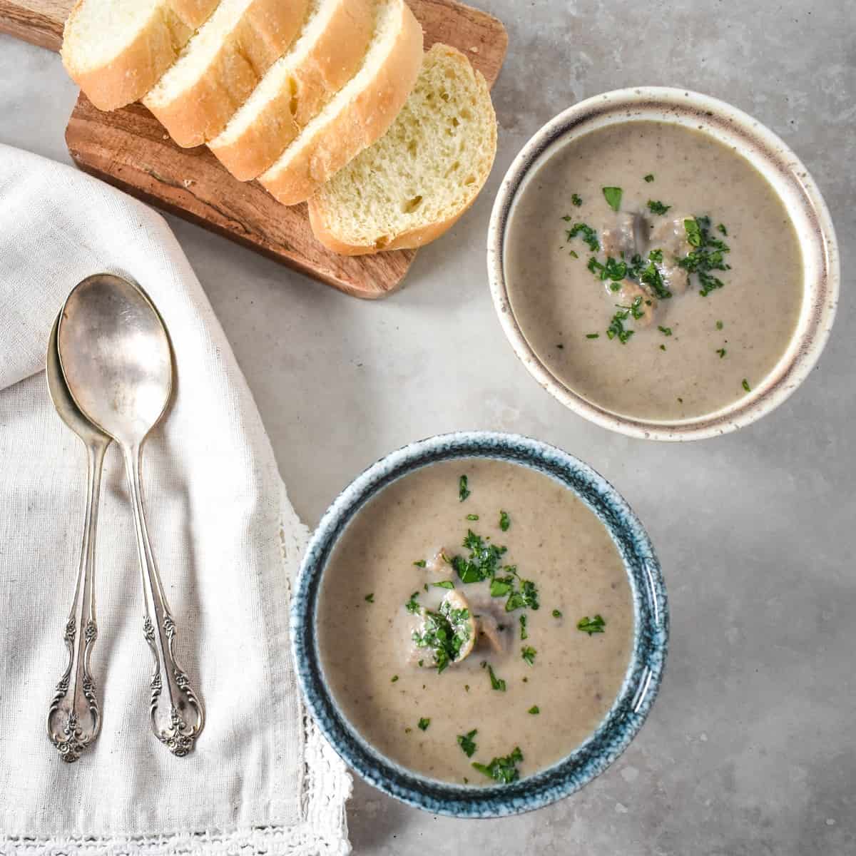 Two small bowls of the mushroom soup one has a blue rim and one a brown rim. They are set on a beige table with sliced bread and spoons on a linen to the left side.