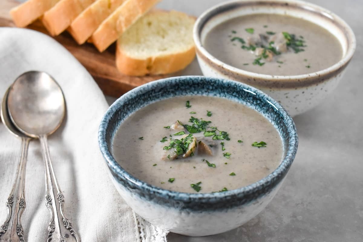 A close up of two small bowls of the mushroom soup one has a blue rim and one a brown rim. They are set on a beige table with sliced bread and spoons on a linen to the left side.