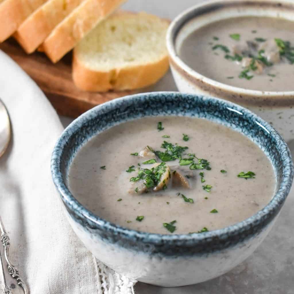 A close up of two small bowls of the mushroom soup one has a blue rim and one a brown rim. They are set on a beige table with sliced bread and spoons on a linen to the left side.