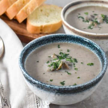 A close up of two small bowls of the mushroom soup one has a blue rim and one a brown rim. They are set on a beige table with sliced bread and spoons on a linen to the left side.