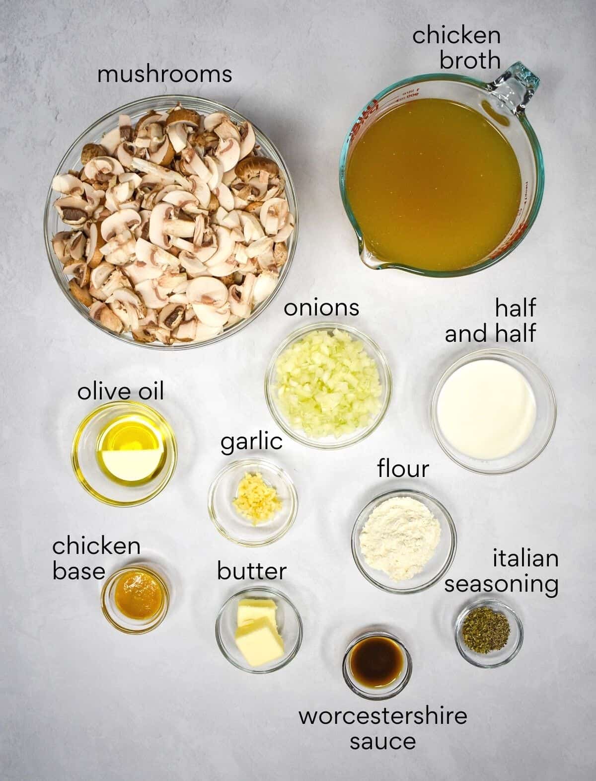 The ingredients for the mushroom soup prepped and arranged in glass bowls on a white table. Each ingredient is labeled with small black letters.
