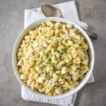 The chicken macaroni salad served in a white bowl, set on a white linen with a serving spoon on the top right side.