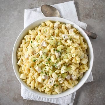 The chicken macaroni salad served in a white bowl, set on a white linen with a serving spoon on the top right side.