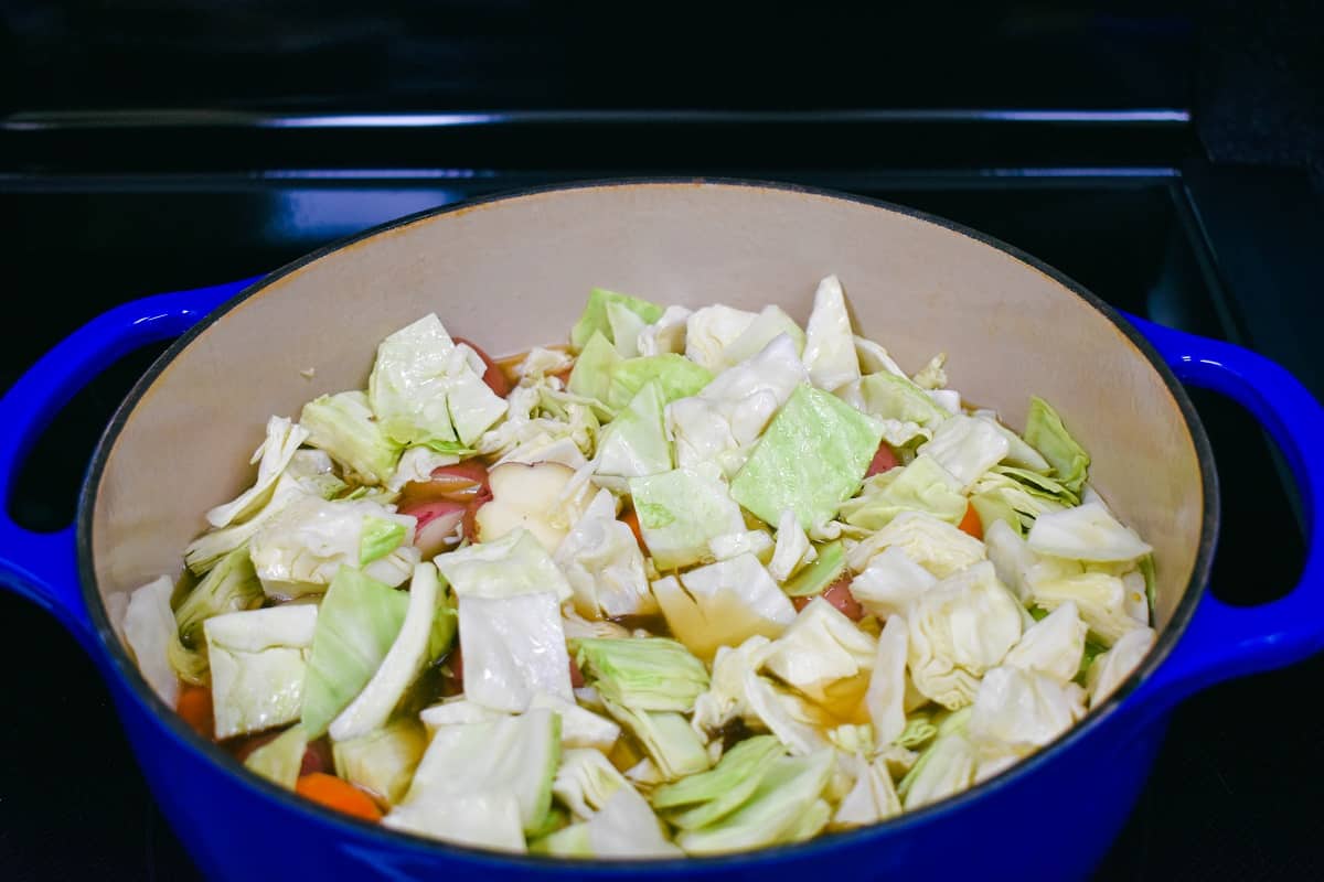 Cabbage, carrots, and potatoes added in broth in a large, blue pot before cooking. 