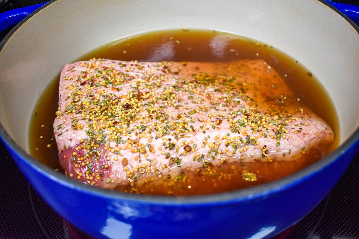 A piece of corned beef with the seasoning on top placed in a large blue pot and almost covered with broth and water.