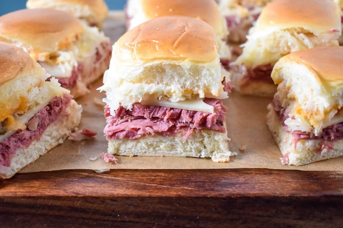 A close up of the prepared corned beef sliders on a piece of parchment paper set on a wood cutting board, after slicing.