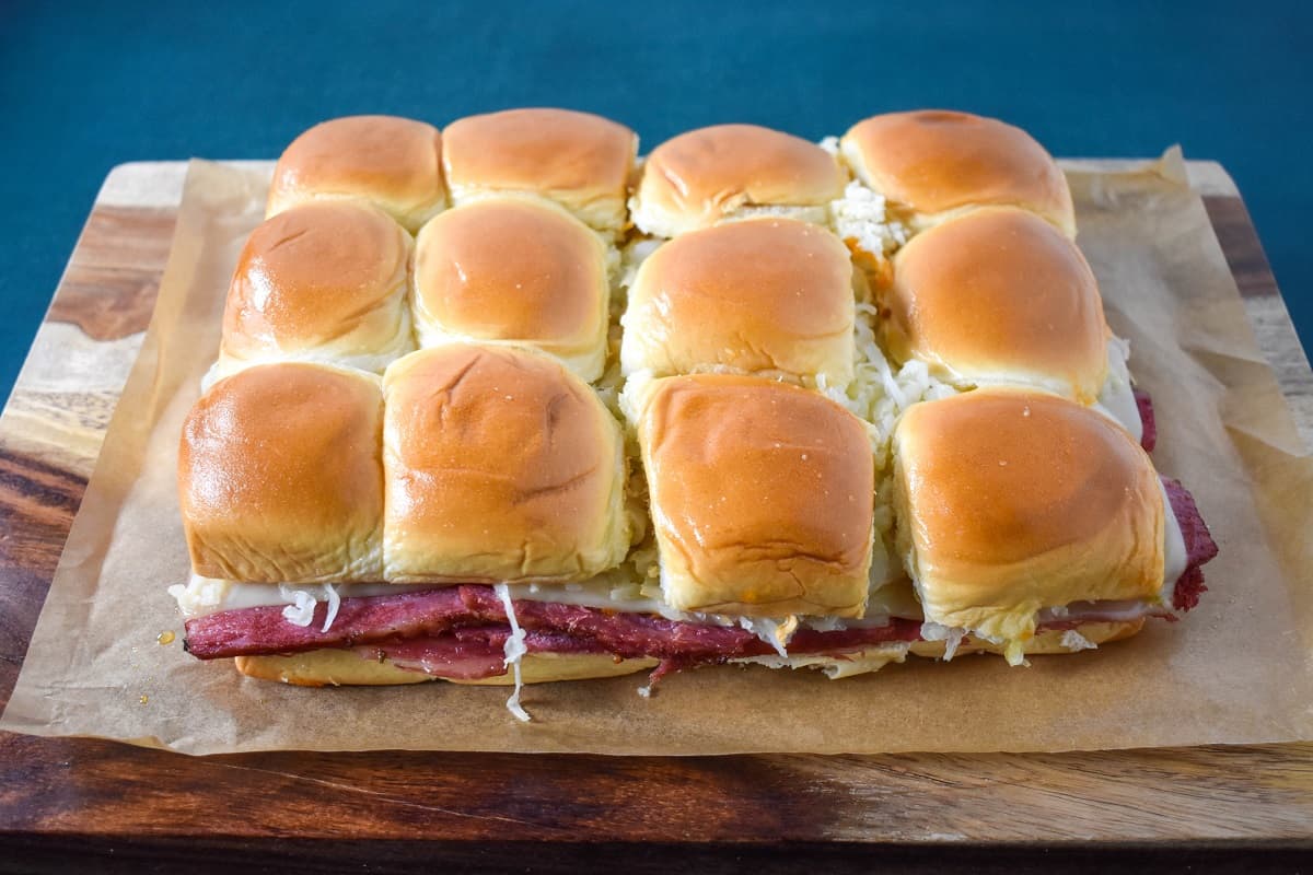 The prepared corned beef sliders on a piece of parchment paper set on a wood cutting board, before slicing.