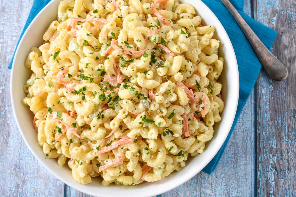 A close-up of the finished macaroni salad garnished with chopped parsley in a large, white serving bowl set on an aqua linen.