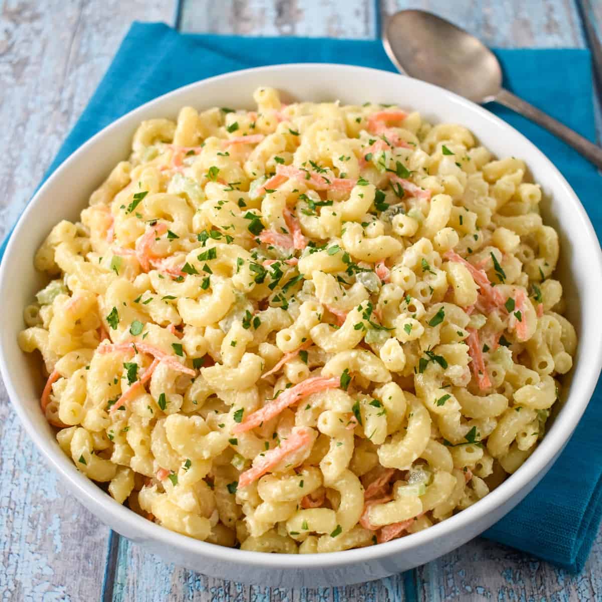 Macaroni salad in a large, white serving bowl set on an aqua linen with a serving spoon in the background.