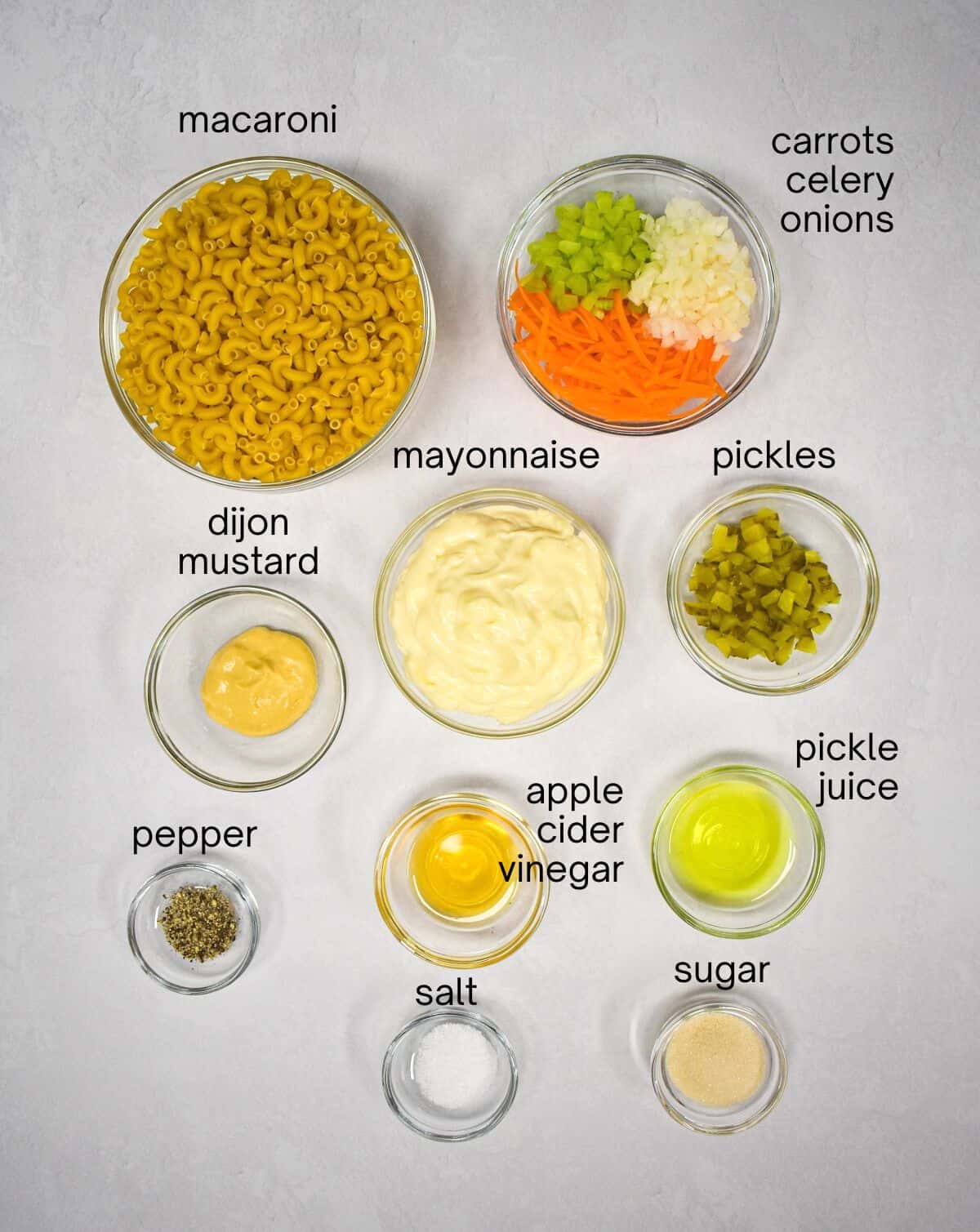 The ingredients for the salad arrange in glass bowls on a white table. Each ingredient is labeled with small black letters.