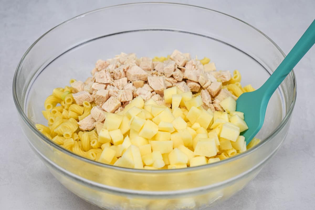 Peeled, diced apple, diced chicken, and macaroni in a large, glass bowl with a teal colored spatula on the right side.