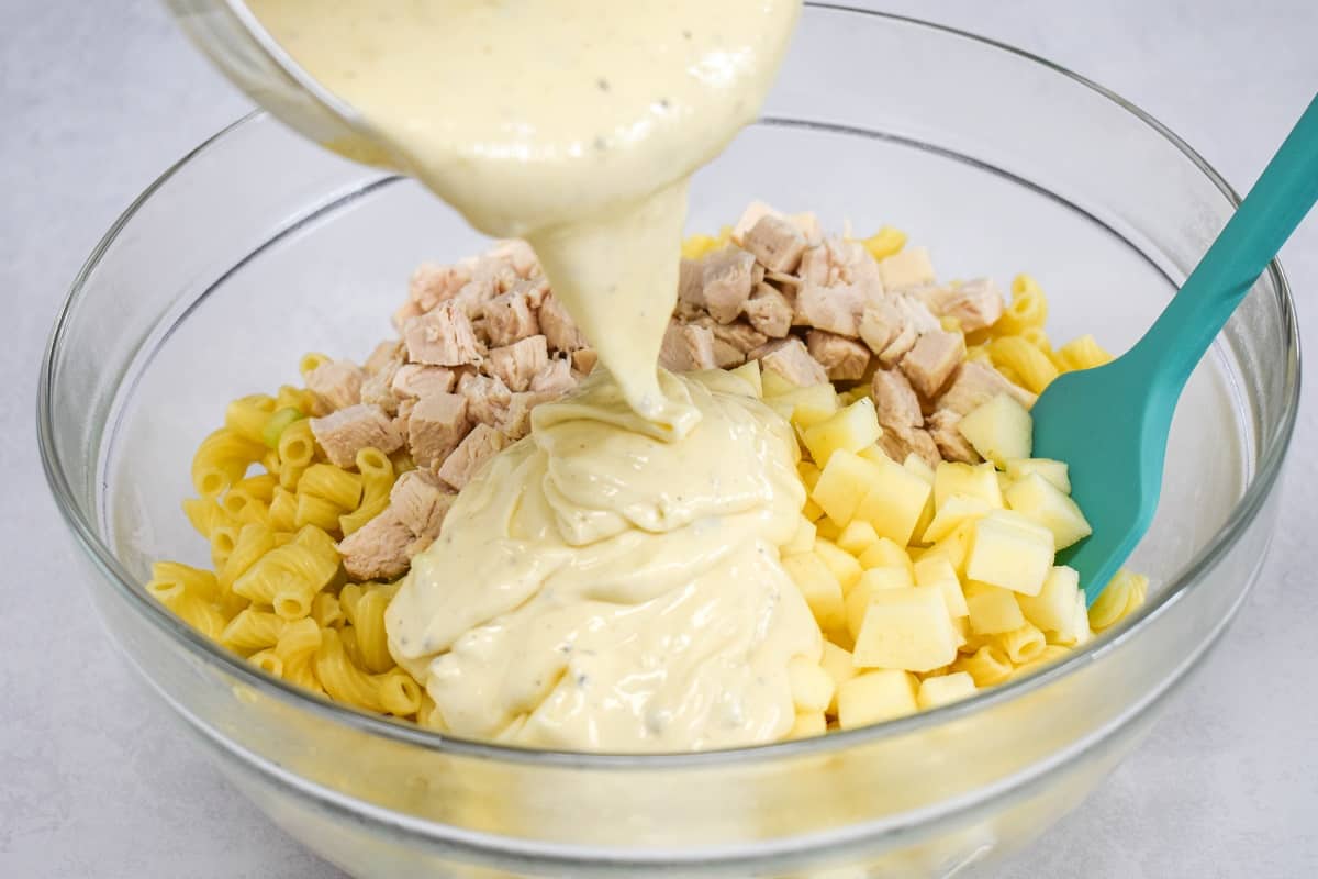The dressing being added to the macaroni and other ingredients in a large, glass bowl. There is a teal colored spatula on the right hand side.