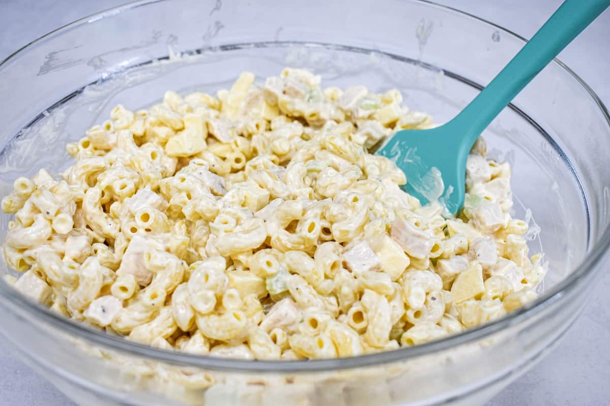 The dressed chicken macaroni salad after being stirred in a large, glass bowl with a teal colored spatula.