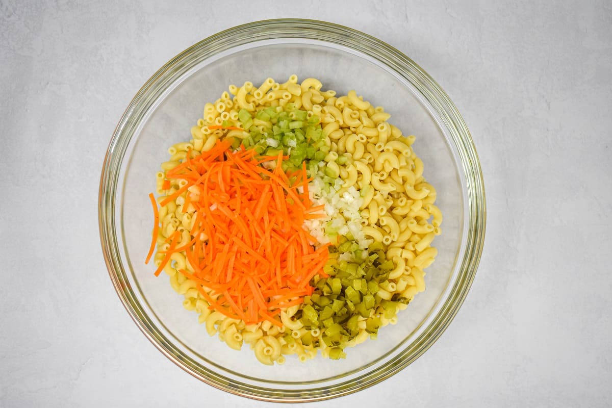 Cooked macaroni, shredded carrots, celery, onions, and pickles in a large, glass bowl.
