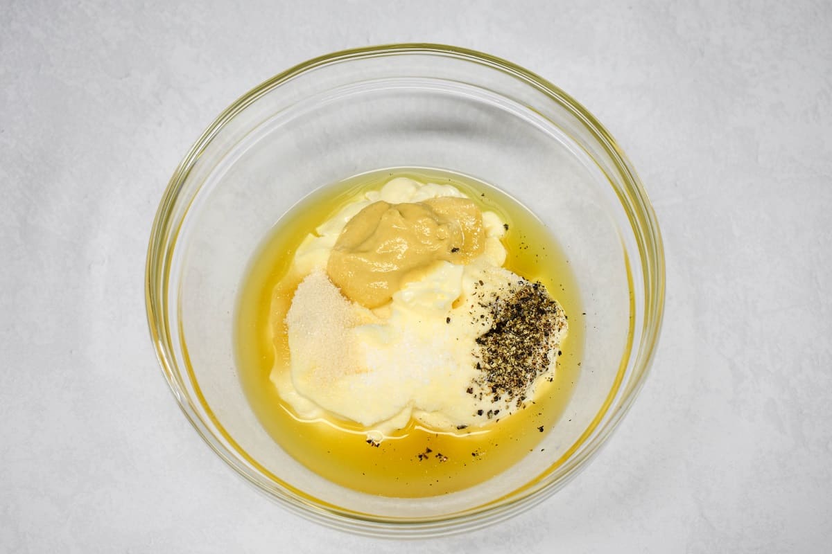 Mayonnaise, dijon mustard, sugar, apple cider vinegar, pickle juice, salt, and pepper in a glass bowl set on a white table.