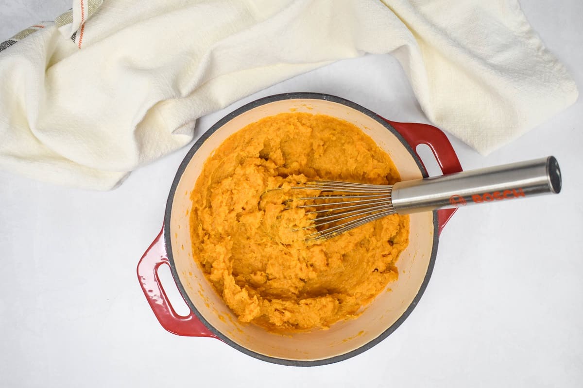 The mashed sweet potato mixture in a red and white pot with a whisk to the right side.